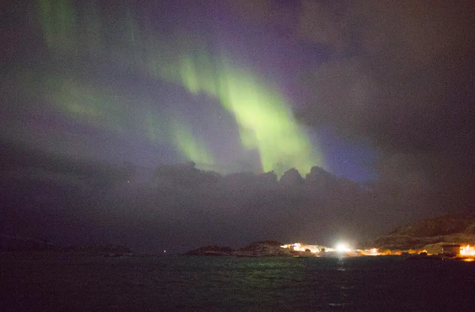 Aurora glowing through thin cloud over the sea at Sommarøy, west of Tromsø, with a few village lights on the shore