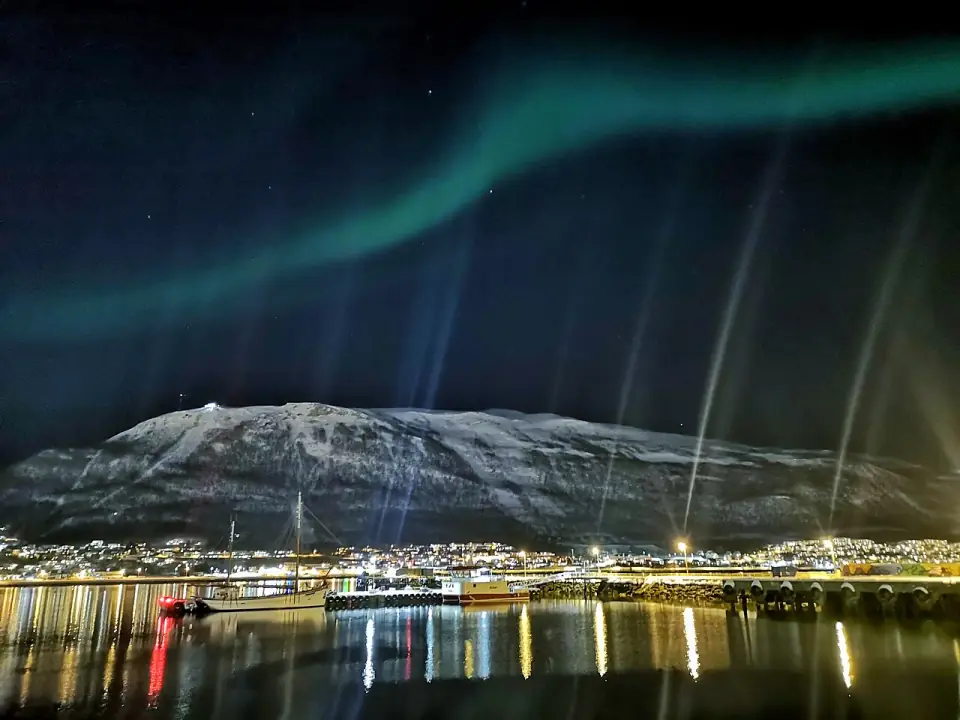 Green aurora over Tromsø at night, the city lights reflected in the sound below the snow-covered mountain